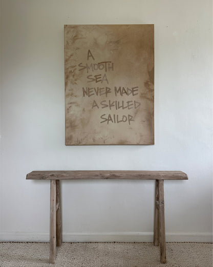 A large, earthy, abstract painting hangs above a rustic vintage wood bench. The neutral, softly textured painting features writing done in sand that reads "A SMOOTH SEA NEVER MADE A SKILLED SAILOR"