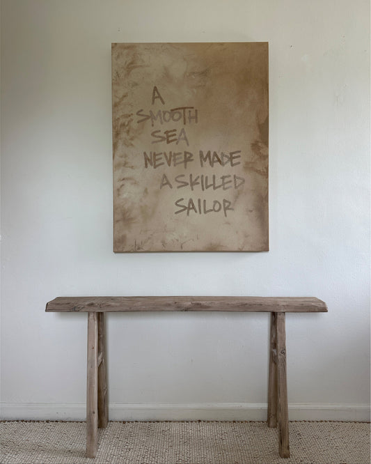 A large, earthy, abstract painting hangs above a rustic vintage wood bench. The neutral, softly textured painting features writing done in sand that reads "A SMOOTH SEA NEVER MADE A SKILLED SAILOR"